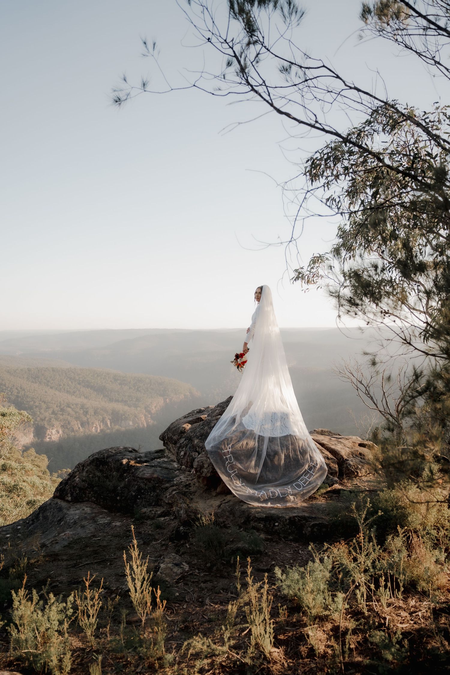 Bride in flowing white gown and long veil stands on rocky cliff overlooking lush mountain valley on her wedding day.