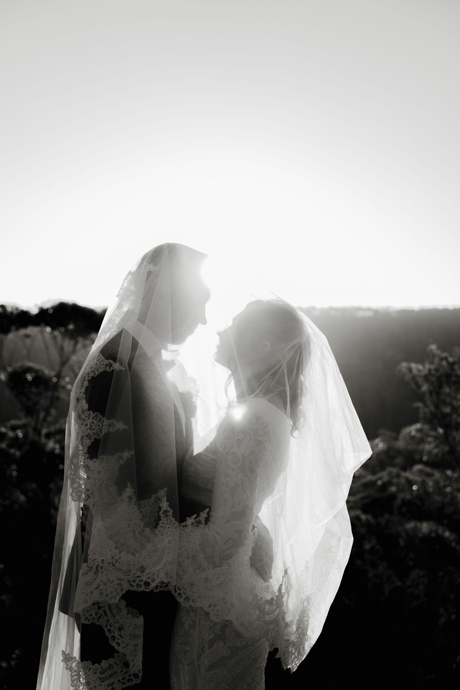 Black and white photo of bride and groom facing each other under veil, backlit by golden sunset light.