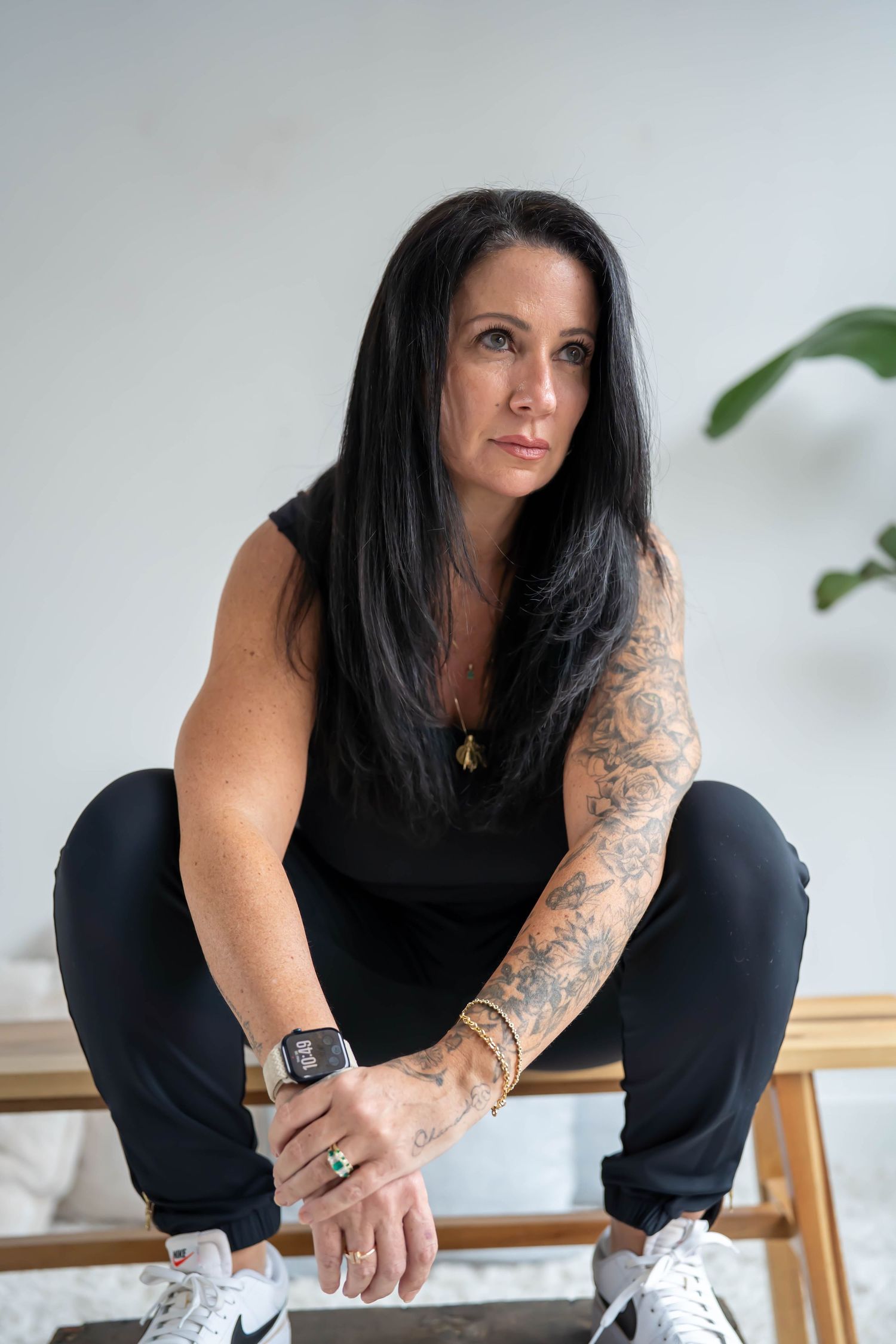 Woman with dark hair and tattoo sleeve in black outfit sits confidently in bright minimalist space.