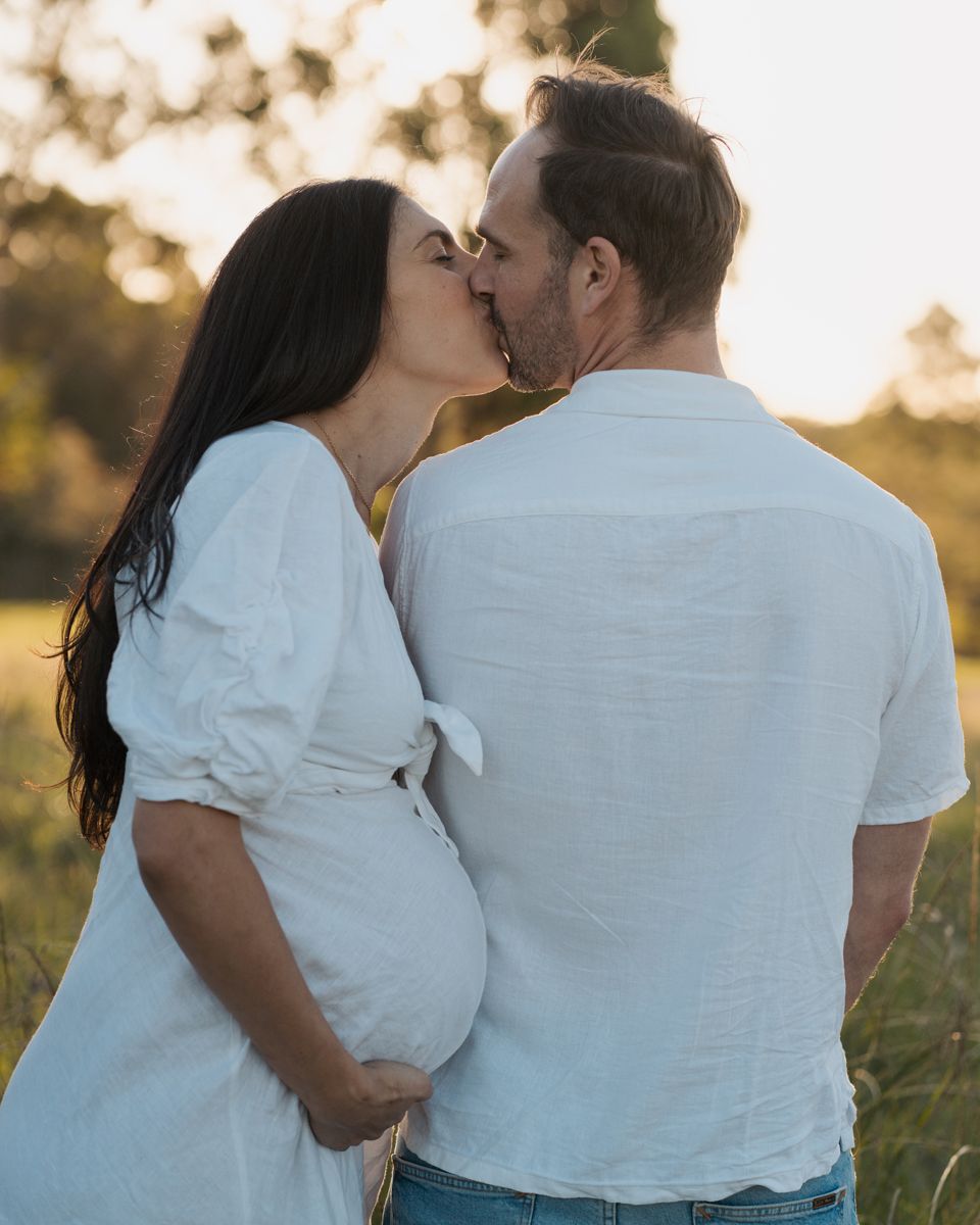Pregnant couple kissing in golden hour sunlight, both wearing white, standing in a green field during maternity photoshoot in rouse hill syd