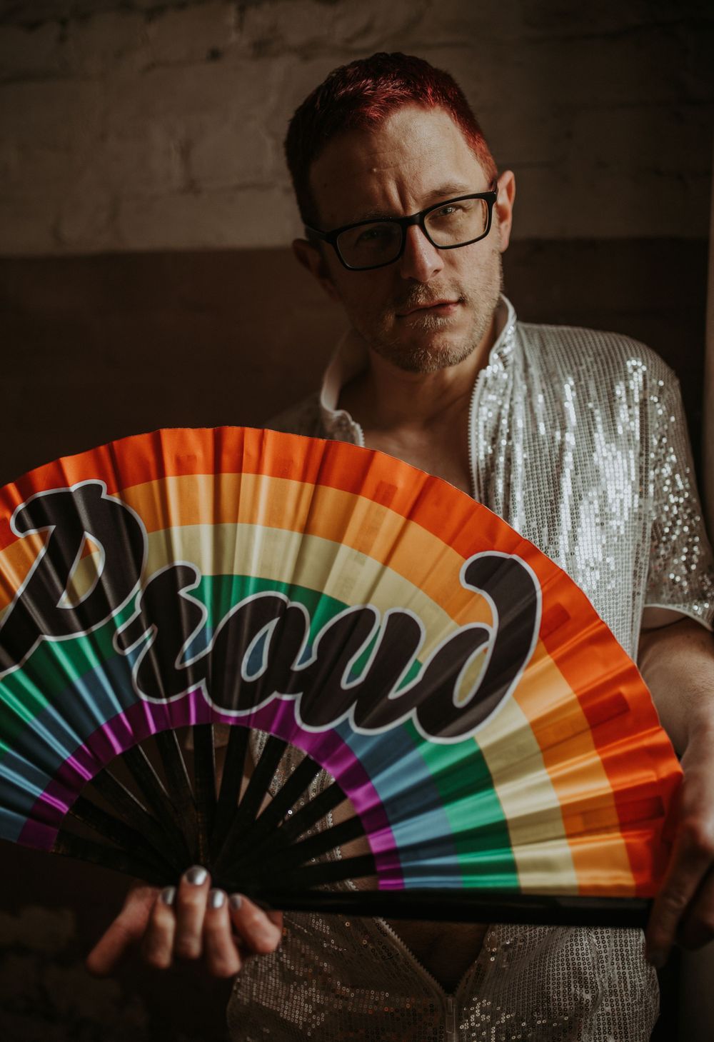 Person holding a rainbow Pride fan with Proud text, wearing silver sequin top with red hair and glasses.