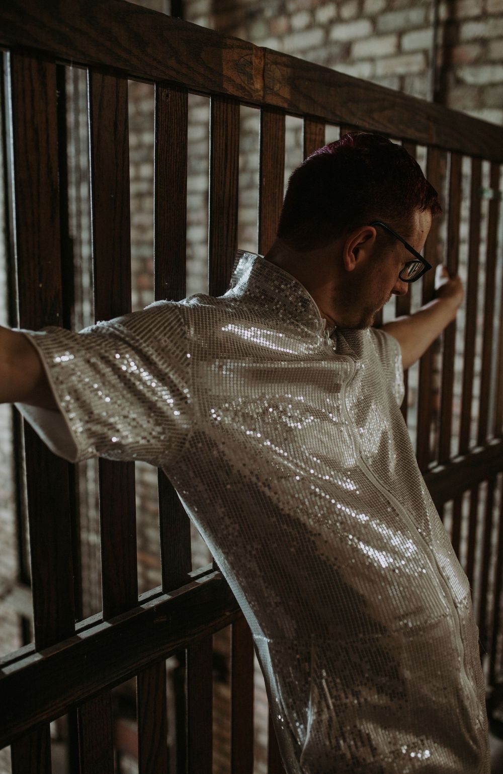 Man in silver sequin shirt leaning against wooden railing with brick wall background.
