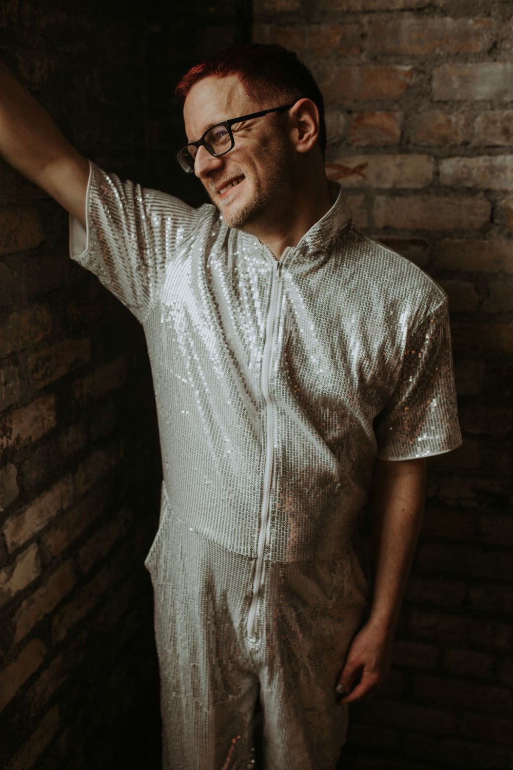 Person in silver sequin jumpsuit leaning against brick wall, smiling with red hair and black glasses.