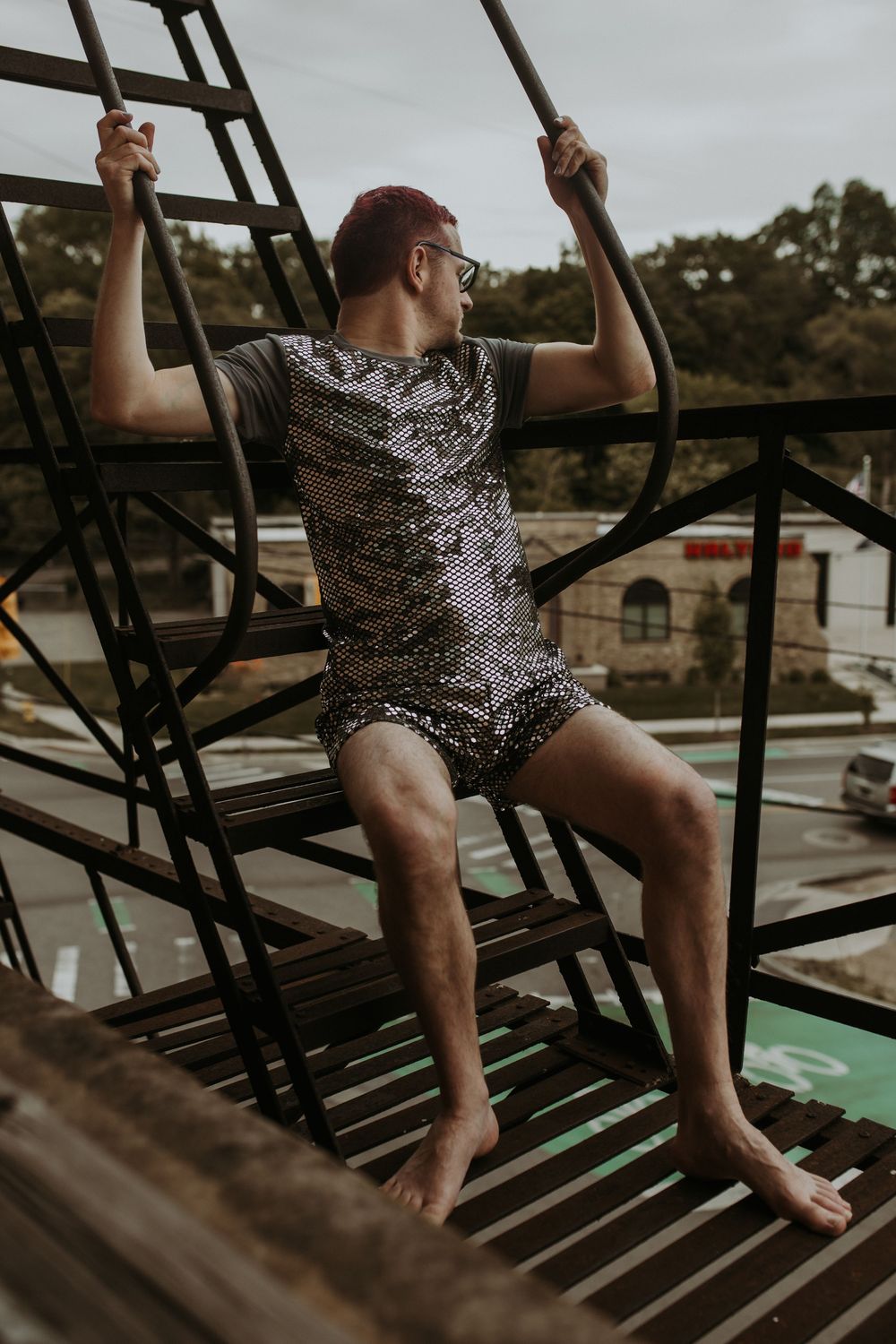 A man in a sequined outfit sits barefoot on metal stairs outdoors, holding rails with trees and street visible behind.