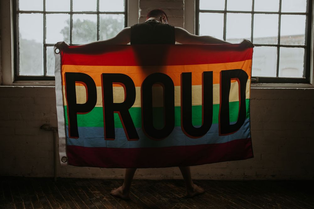 Person holding a rainbow Pride flag with PROUD text in a sunlit room, celebrating LGBTQ+ pride.