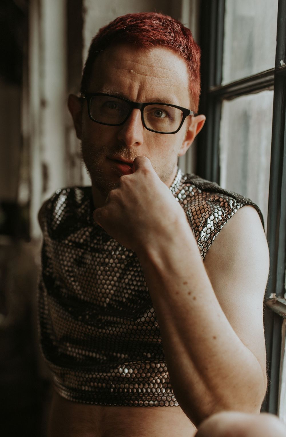 Person with red hair and glasses wearing gold sequin crop top, posing thoughtfully near window.