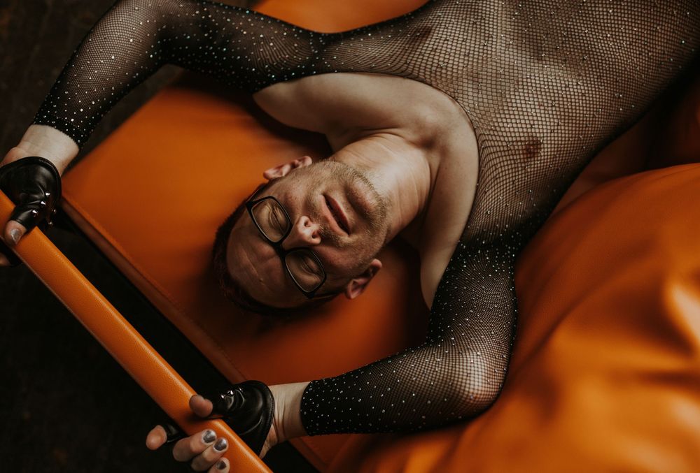 A person in a sparkly black mesh bodysuit lies back on an orange surface, head tilted back in an artistic pose.