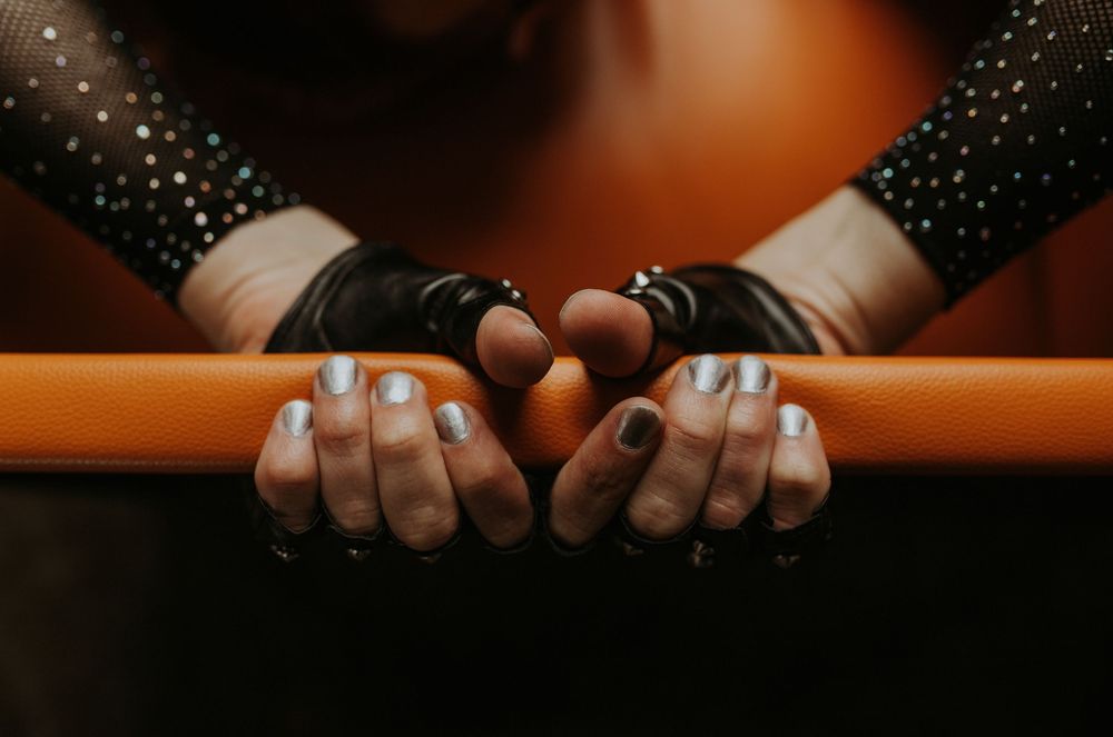 Close-up of gymnast hands gripping an orange balance beam, wearing black grips with iridescent manicured nails.