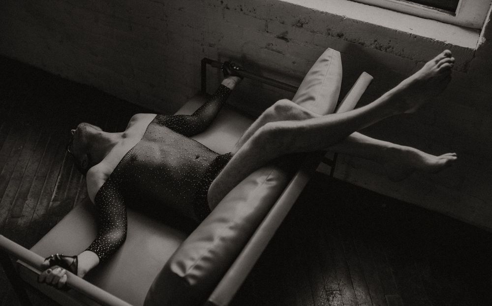 Black and white photo of a person reclining on a vintage chaise lounge near a window on a wooden floor.