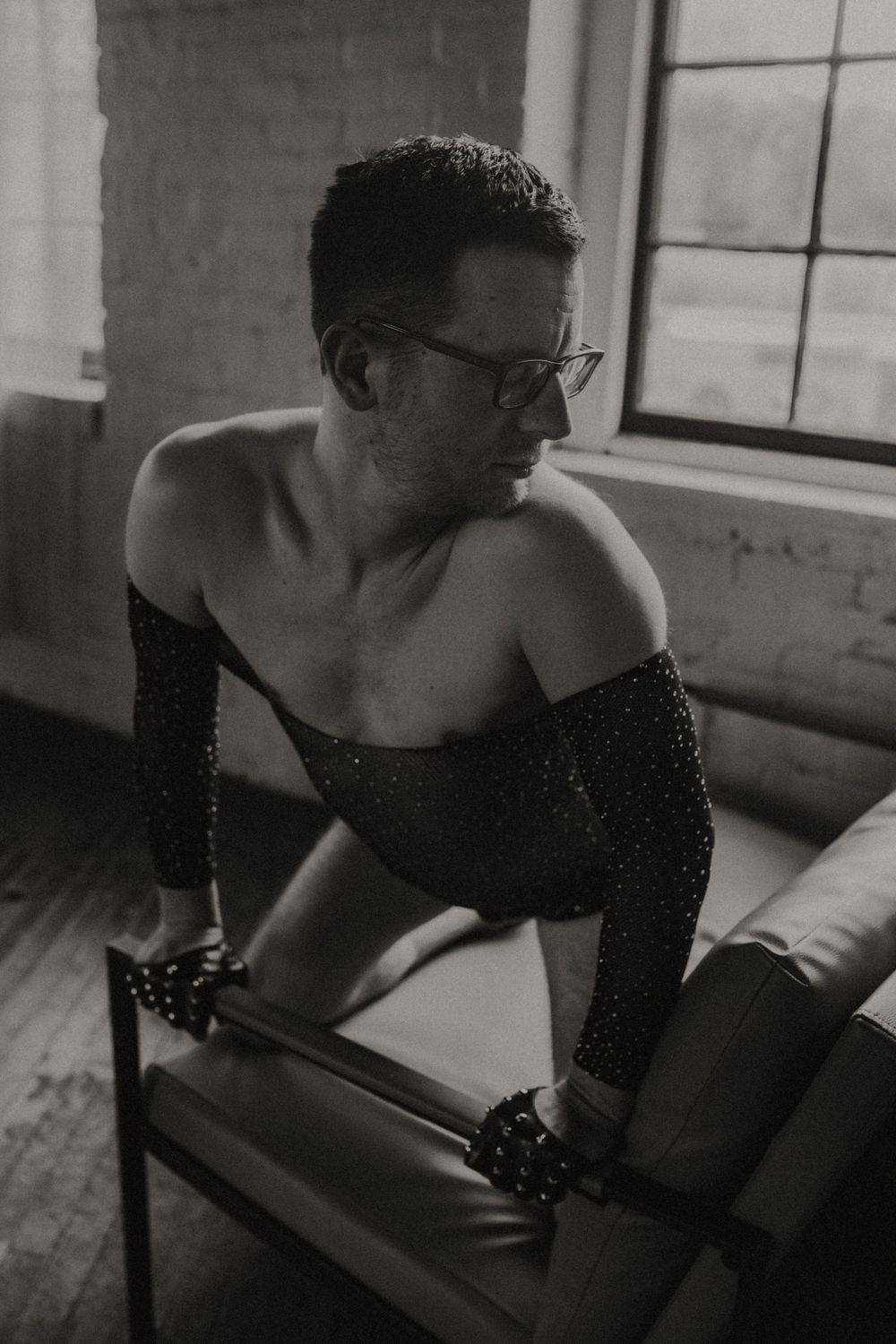 Black and white photo of a shirtless person in sparkly sleeves and glasses leaning on a chair near windows.