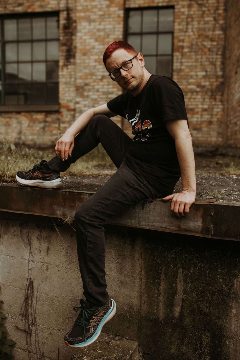 Man with red hair and glasses sitting on concrete ledge wearing black t-shirt and sneakers near brick building.