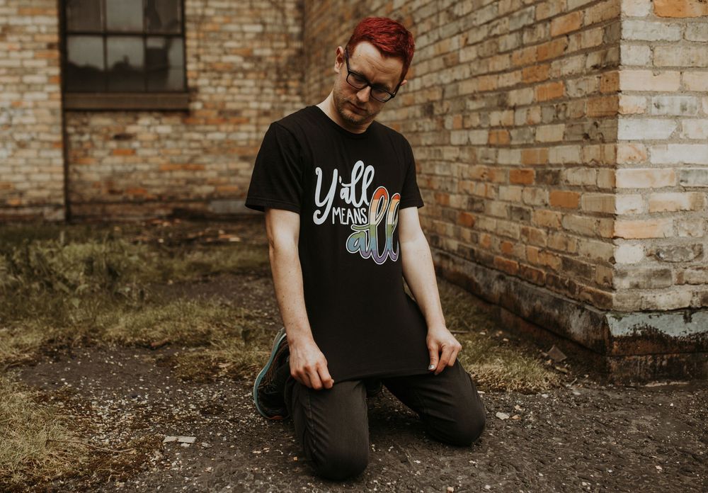 Person kneeling outdoors in black graphic tee reading 'y'all means all' near brick building.