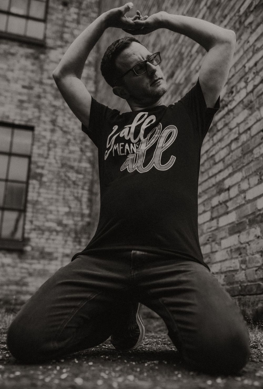 Man kneeling outdoors in black graphic t-shirt reading 'all means all' with hands raised above head.