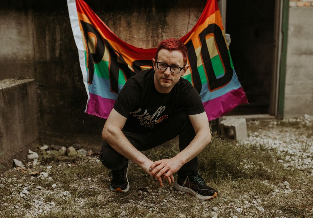 Man crouching in front of a colorful Pride rainbow flag in an outdoor urban setting.