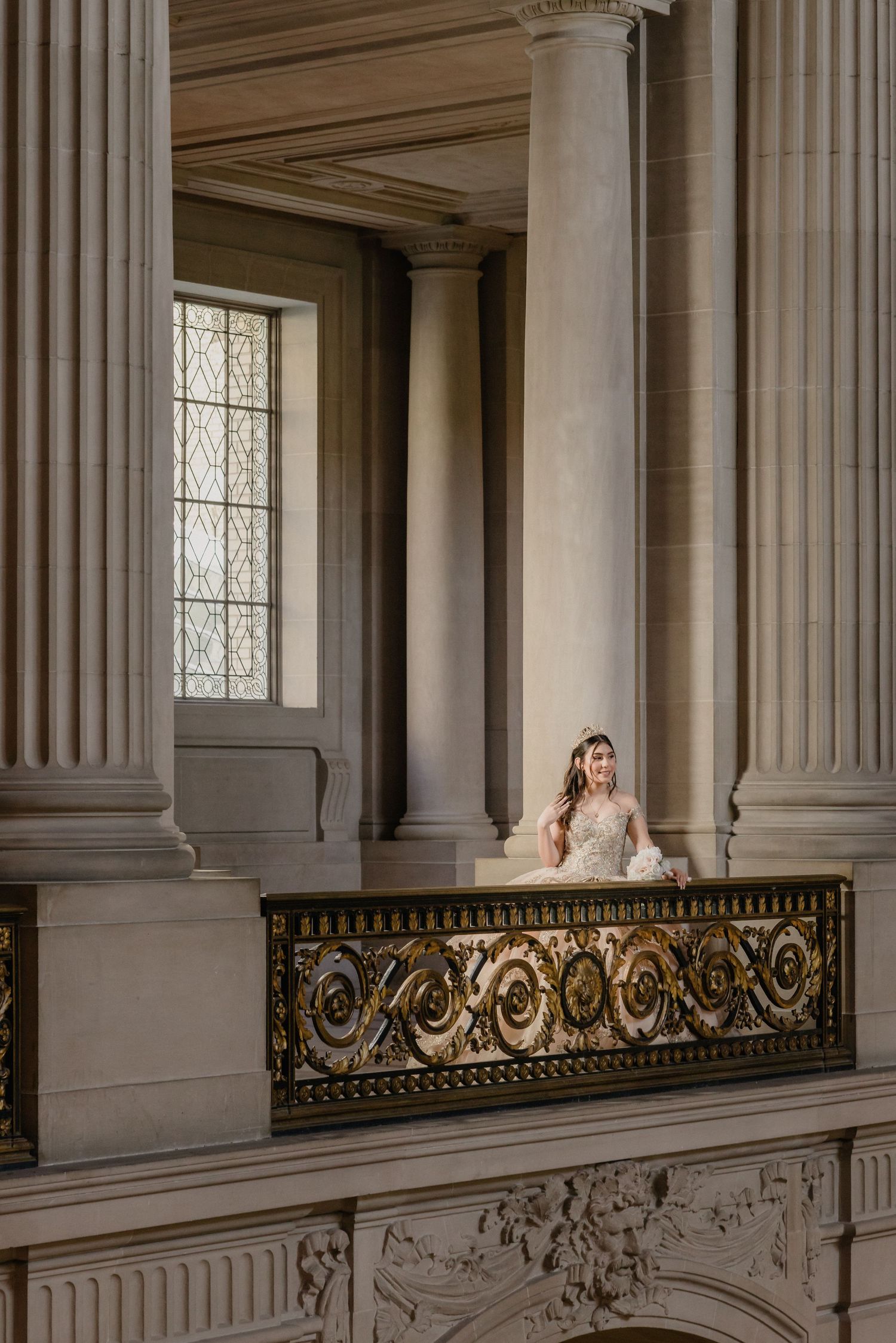 Bride in wedding gown seated on ornate balcony with gold railing amid grand neoclassical columns.