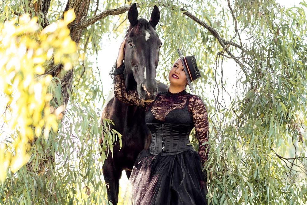 Woman in gothic black corset dress poses with a black horse under weeping willow trees portrait photoshoot with a horse in sydney