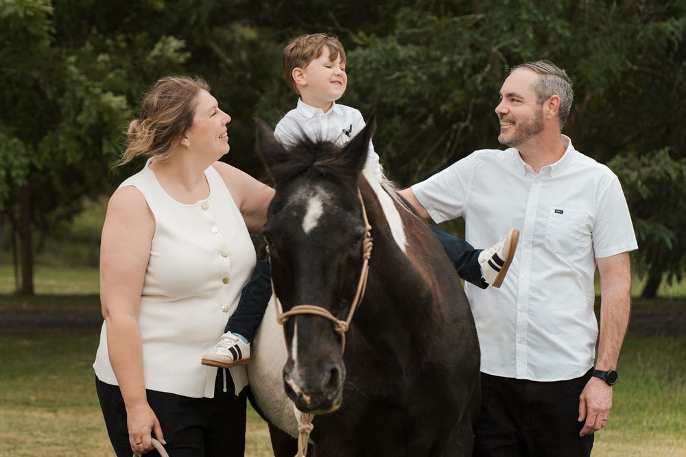 Family of three smiling outdoors with a black and white horse on a green lawn during a photoshoot with a horse in sydney