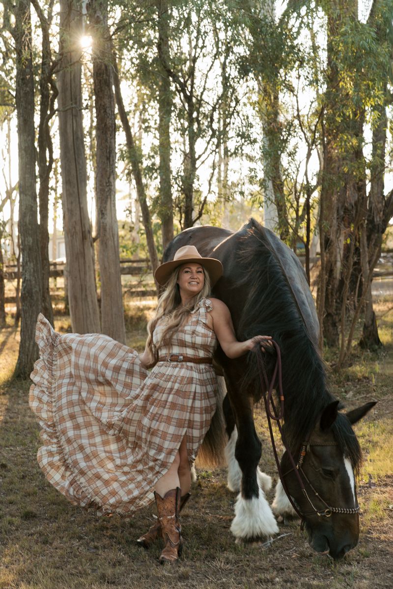 Woman in plaid dress and cowboy hat poses with dark horse in sunlit forest setting during portrait photoshoot in sydney with a horse