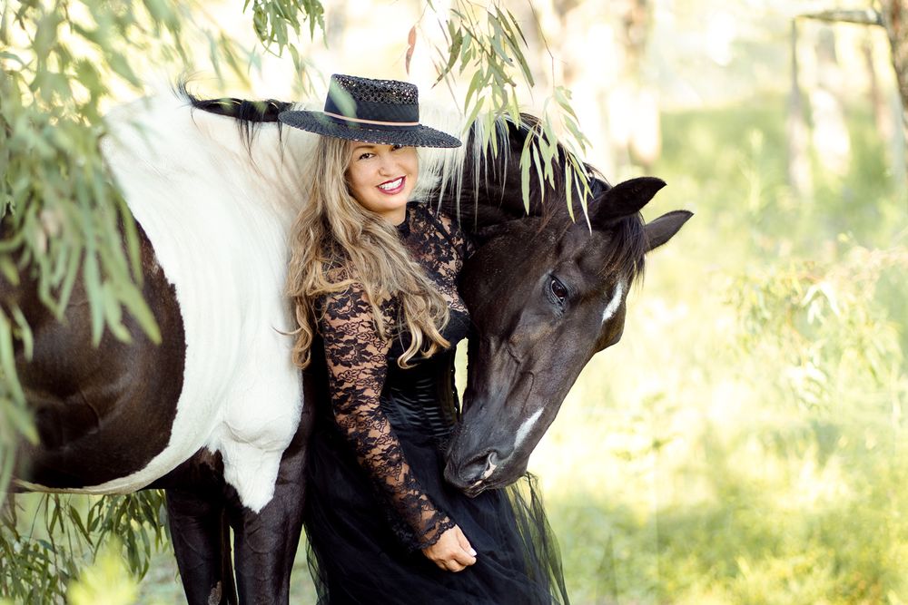 Woman in black lace dress and wide-brim hat posing with a dark horse among green trees during a photoshoot with a horse in sydney