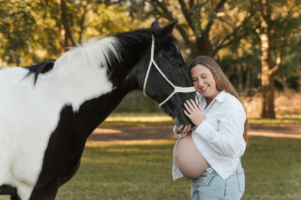Pregnant woman in white shirt smiling with black and white horse outdoors in green park setting maternity horse photoshoot in sydney