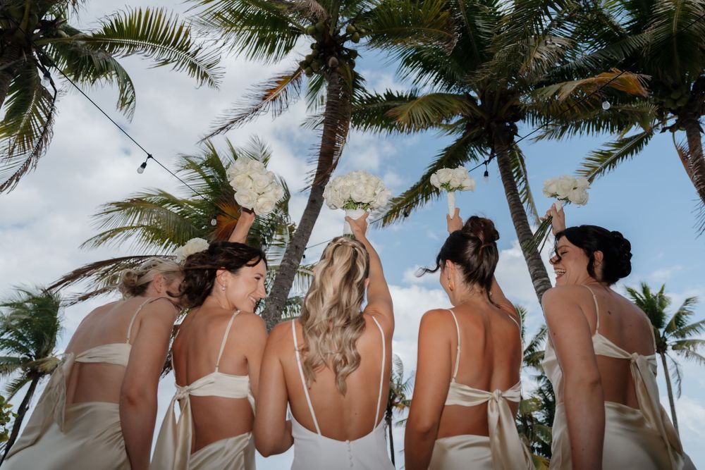 Bride and bridesmaids celebrating with bouquets surrounded by tropical palms during getting ready in Riviera Maya wedding photography.