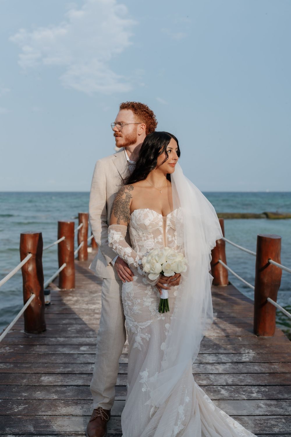 Bride and groom pose on a wooden pier by the ocean, she holds white roses bouquet at The Fives Beach Hotel & Residences.