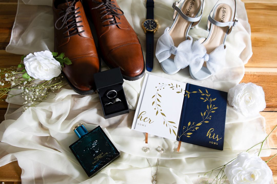 Wedding flat lay with brown dress shoes, navy watch, ring box, his and her vow books, heels, perfume, and white florals.