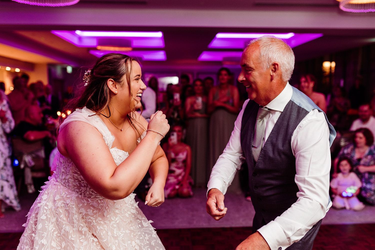 Bride and older man share a joyful father-daughter dance at a wedding reception with guests watching.