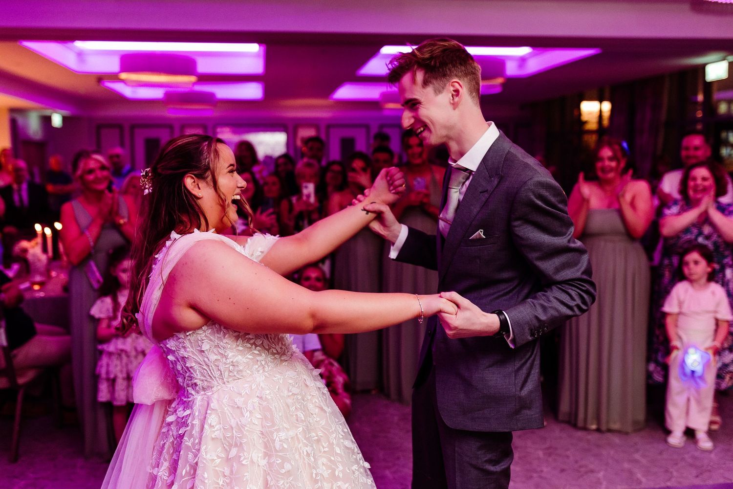 Bride and groom share their first dance at a wedding reception with guests applauding in the background.