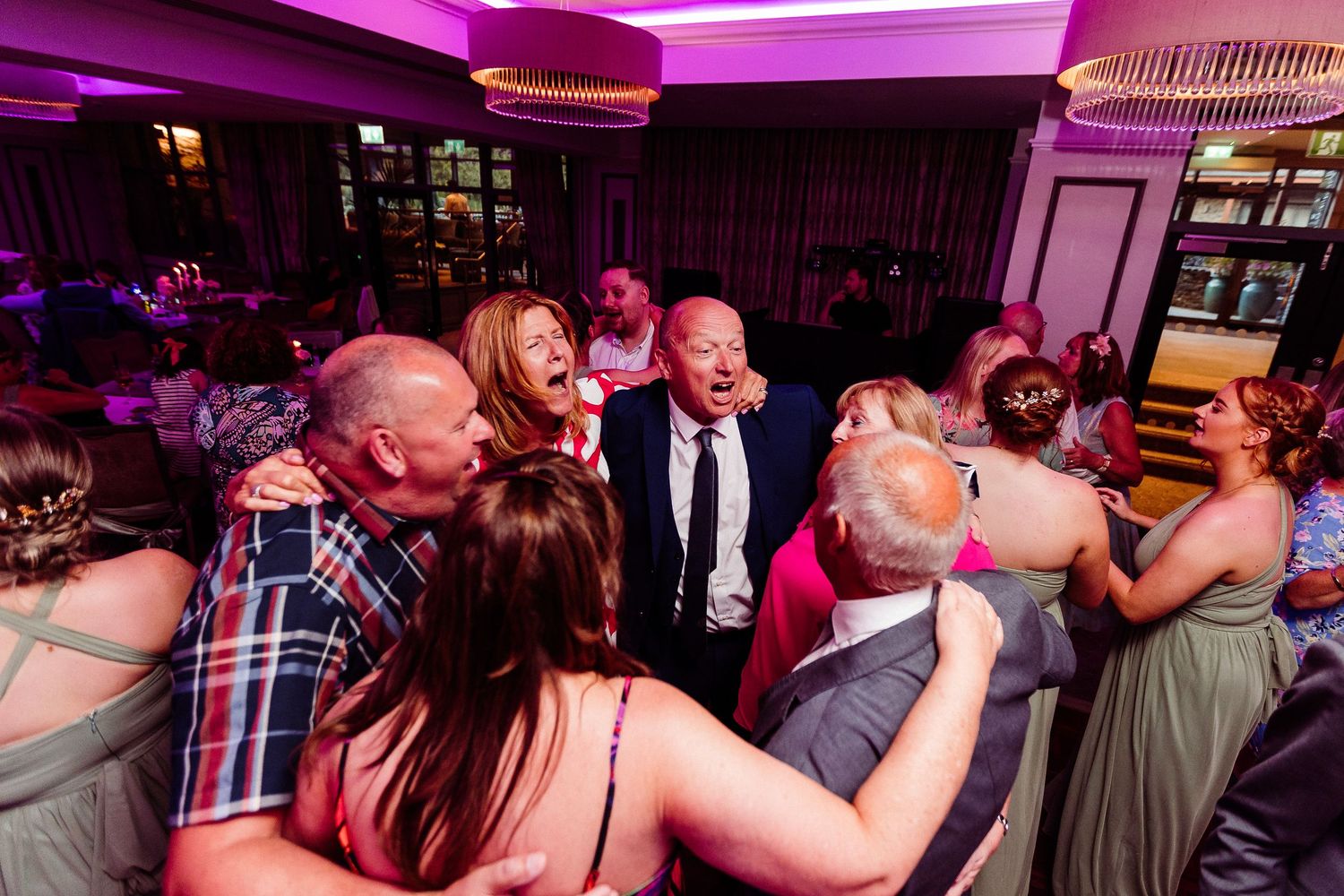 Guests laughing and dancing together in a lively wedding reception under purple lights.
