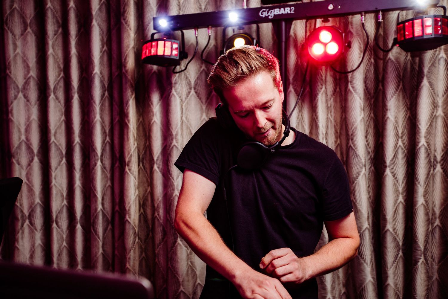 A DJ in a black t-shirt performs under colorful stage lights in a dimly lit venue.