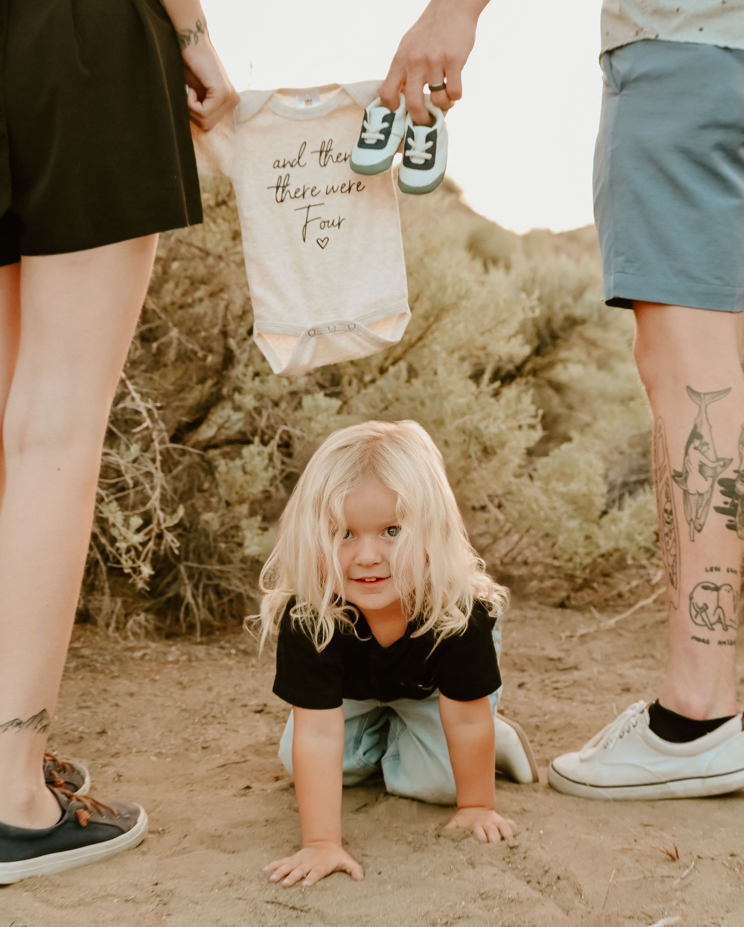 Family pregnancy announcement with toddler holding baby onesie and tiny shoes outdoors in desert setting.