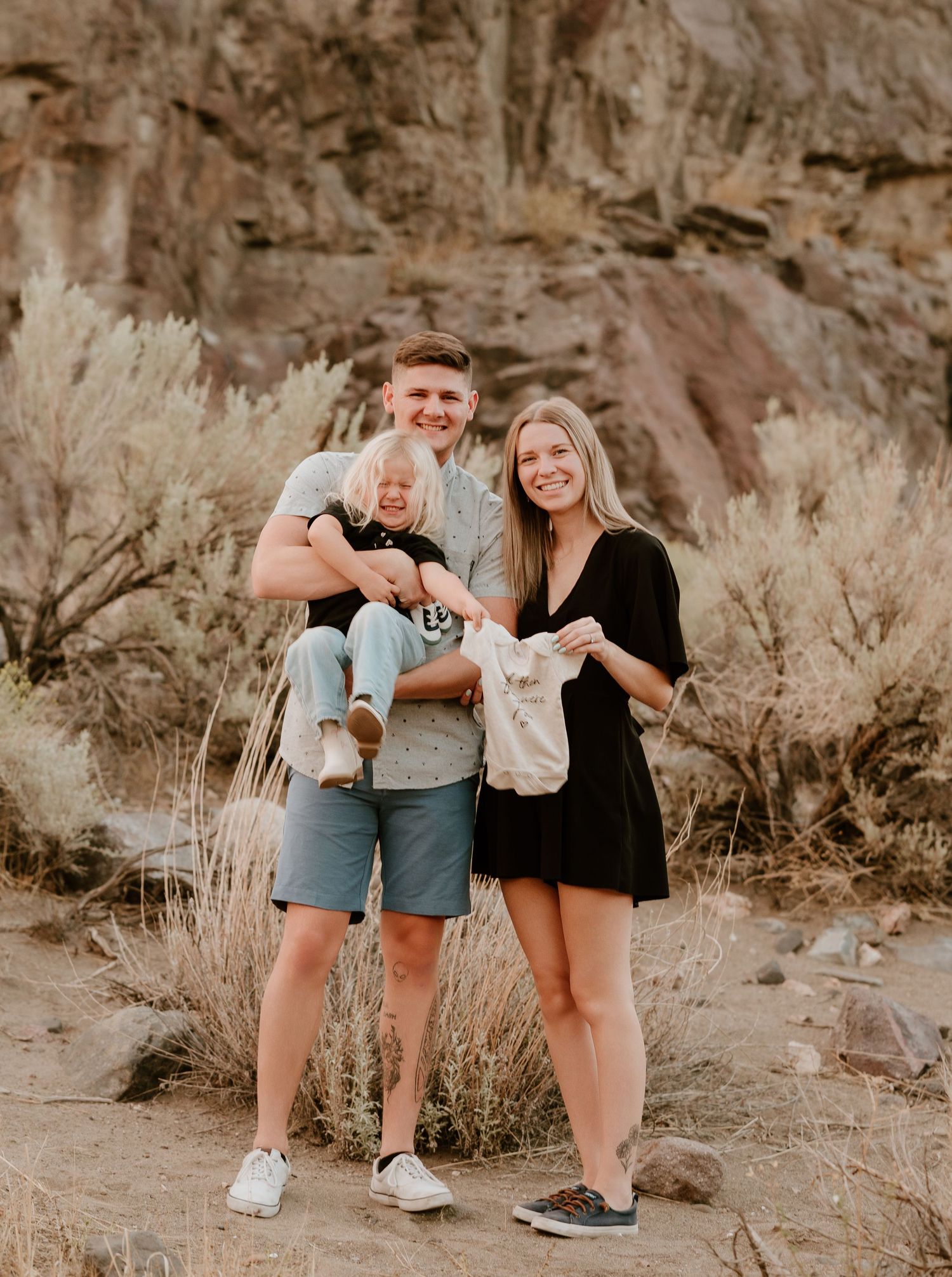 A young family poses outdoors in a desert setting, man holding toddler, woman in black dress, rocky cliffs behind them.