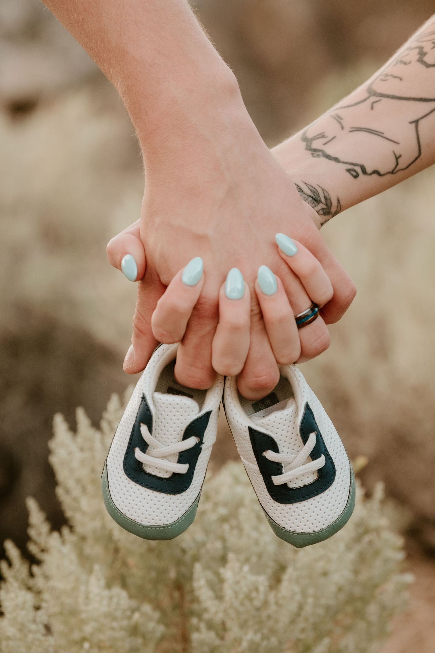 Two hands holding tiny baby sneakers outdoors, one with mint nails and a ring, the other with a tattoo.