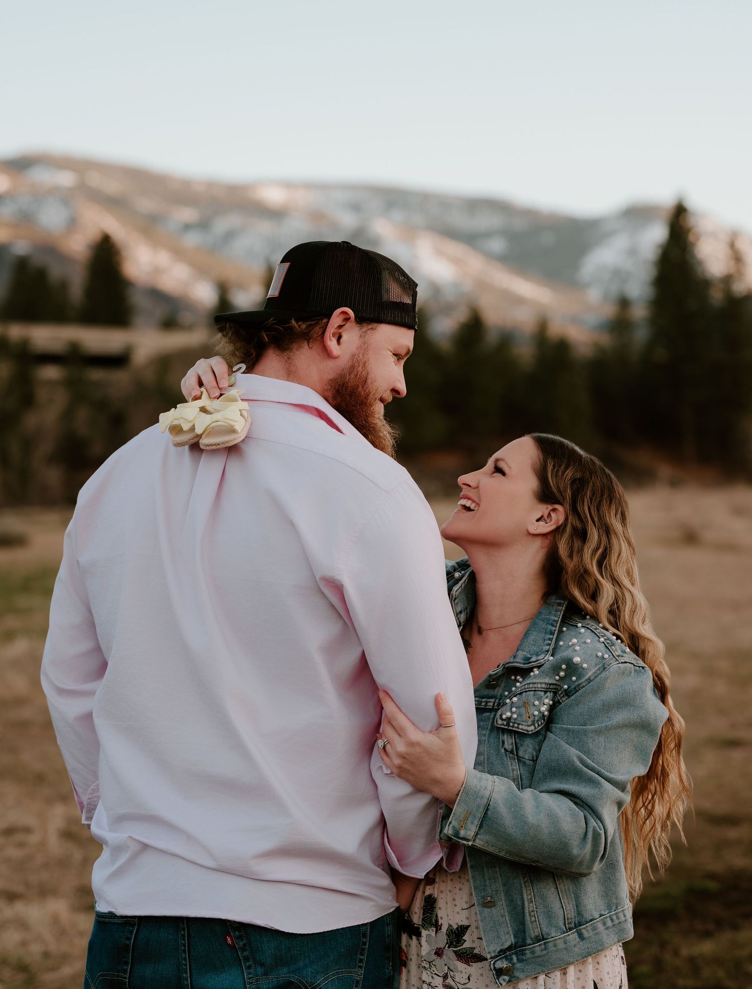 Couple embracing in a mountain meadow at sunset, woman in denim jacket smiling at bearded man in pink shirt.