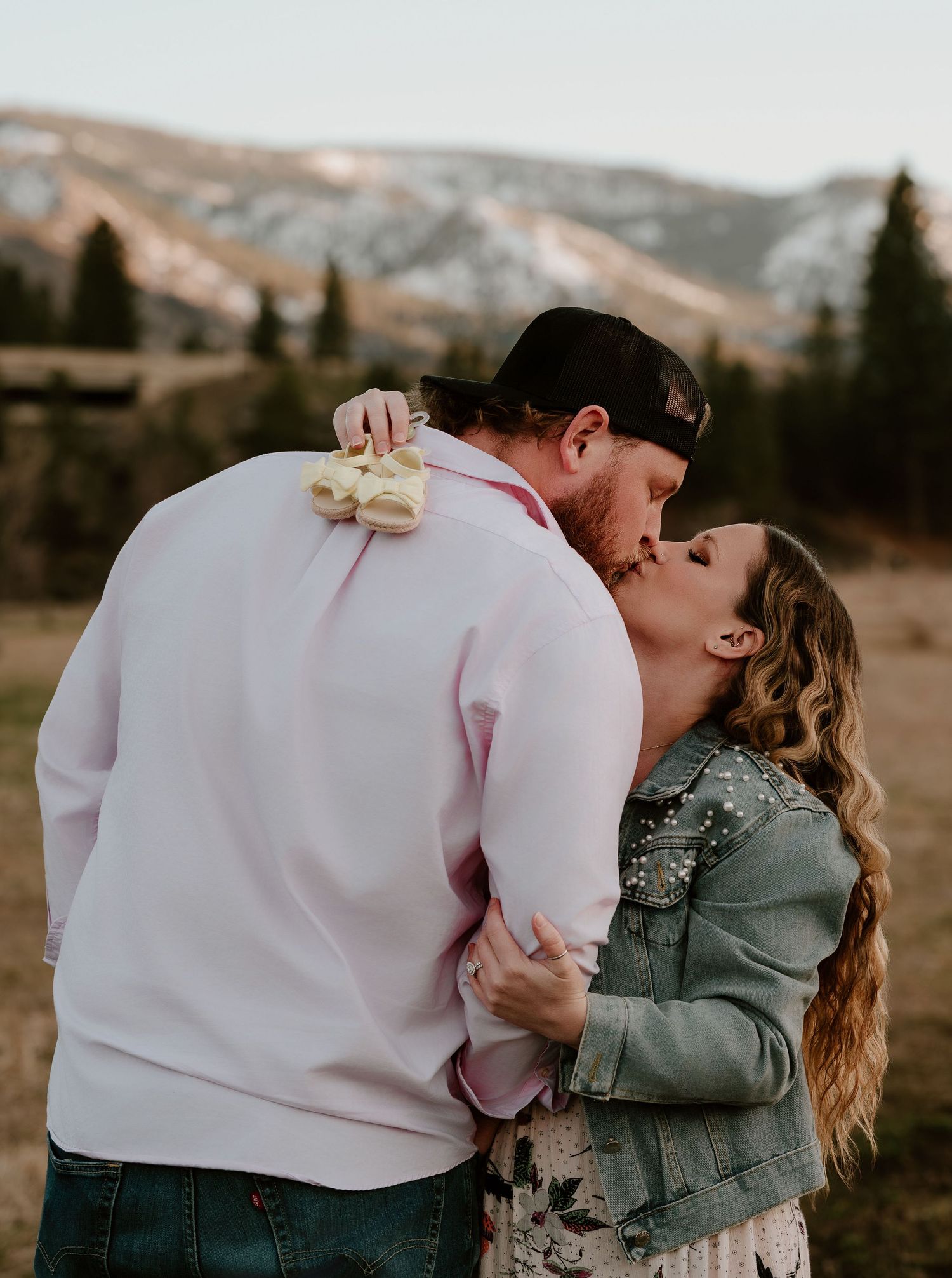 Couple sharing a romantic kiss outdoors holding tiny baby shoes, with mountain scenery in background.