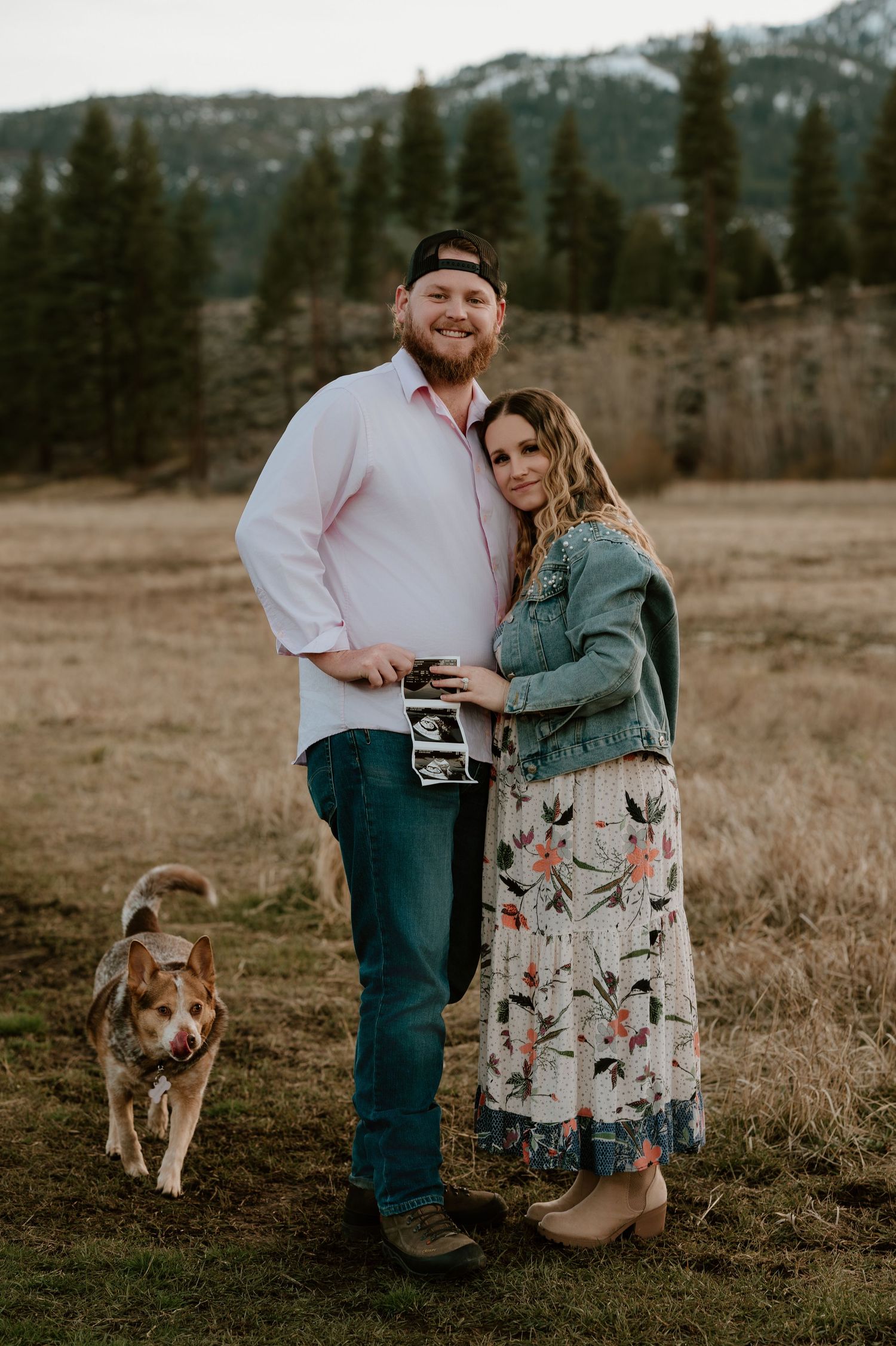 Couple holding ultrasound photo in mountain meadow with pine trees, dog nearby, pregnancy announcement outdoor photo.