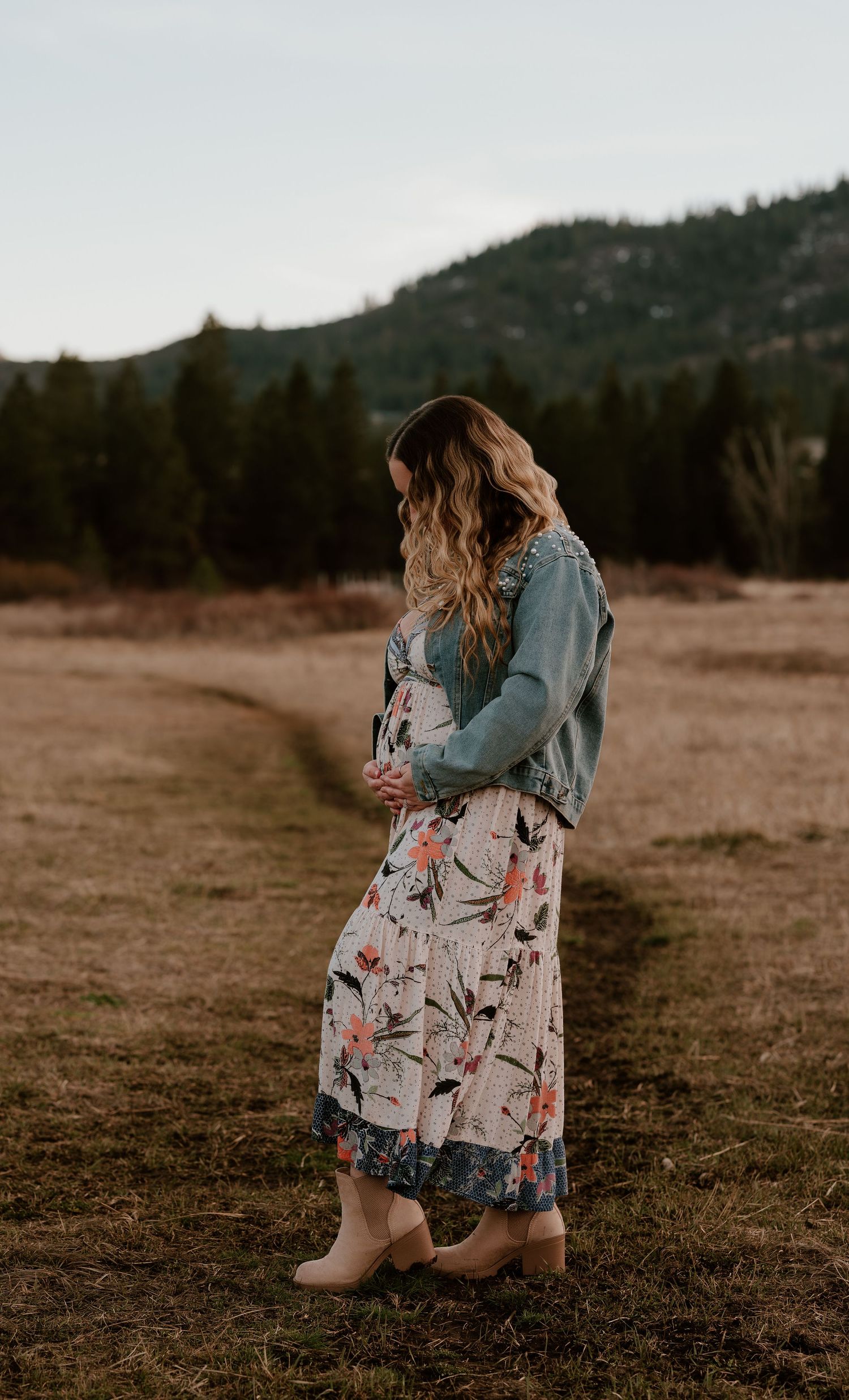 Pregnant woman in floral dress and denim jacket standing in a field with pine trees and mountains in background.