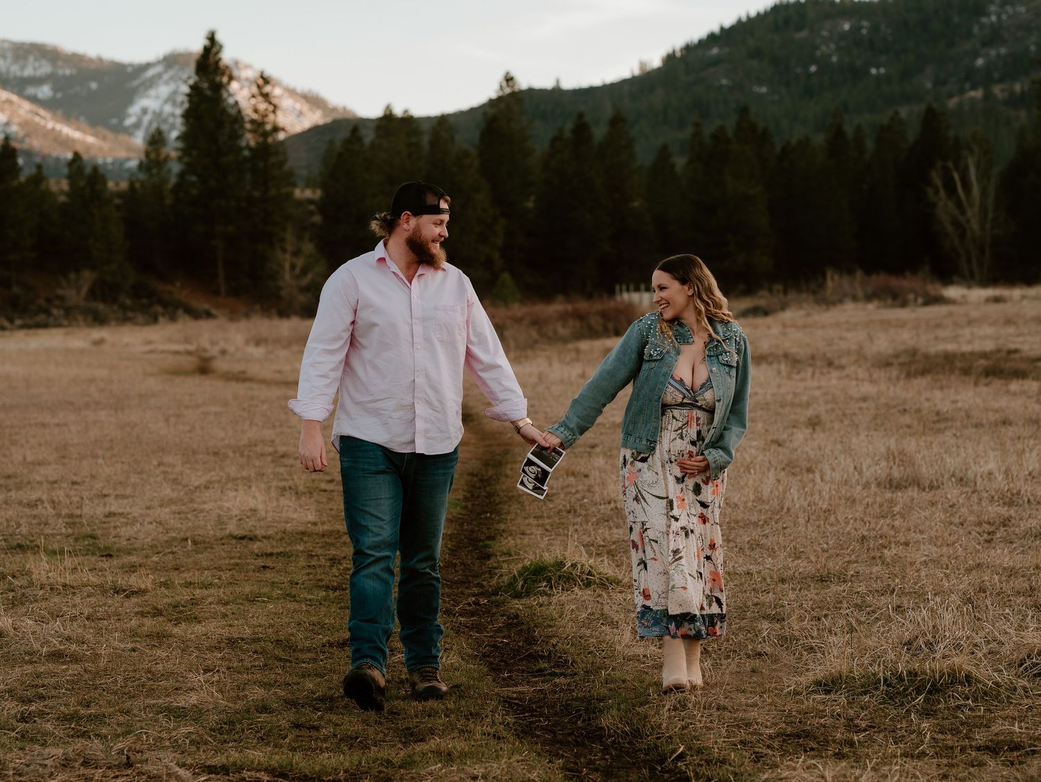 Couple holding hands walking through a mountain meadow with pine trees, man in pink shirt and woman in floral dress.