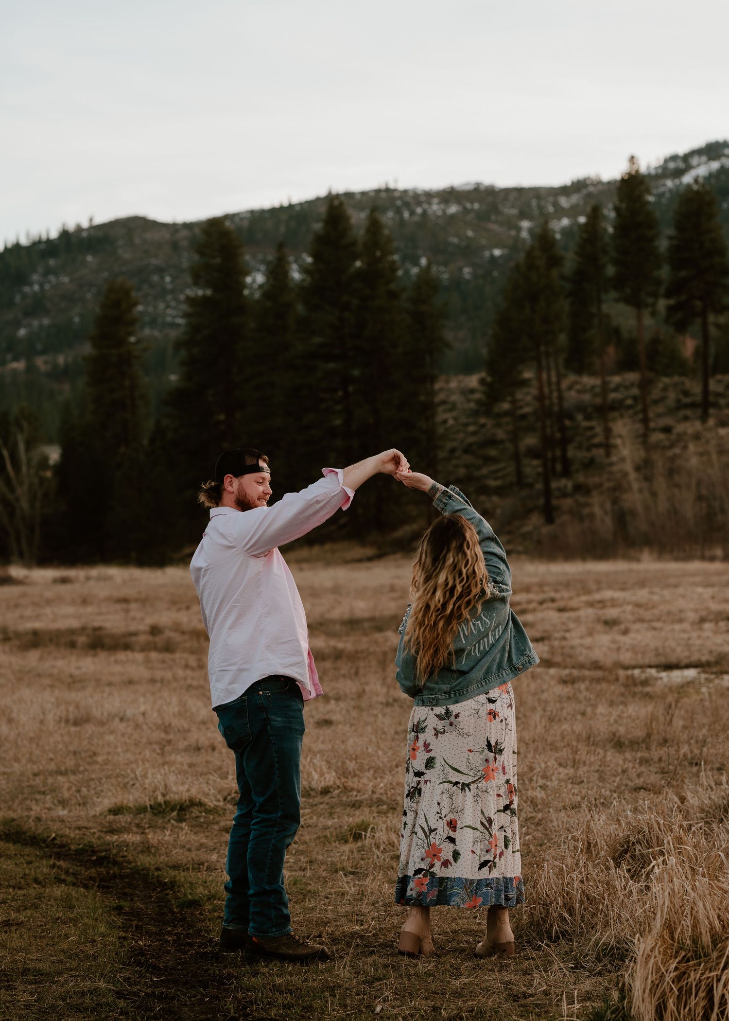 Couple dancing together in a mountain meadow surrounded by pine trees, woman spinning in floral dress and denim jacket.