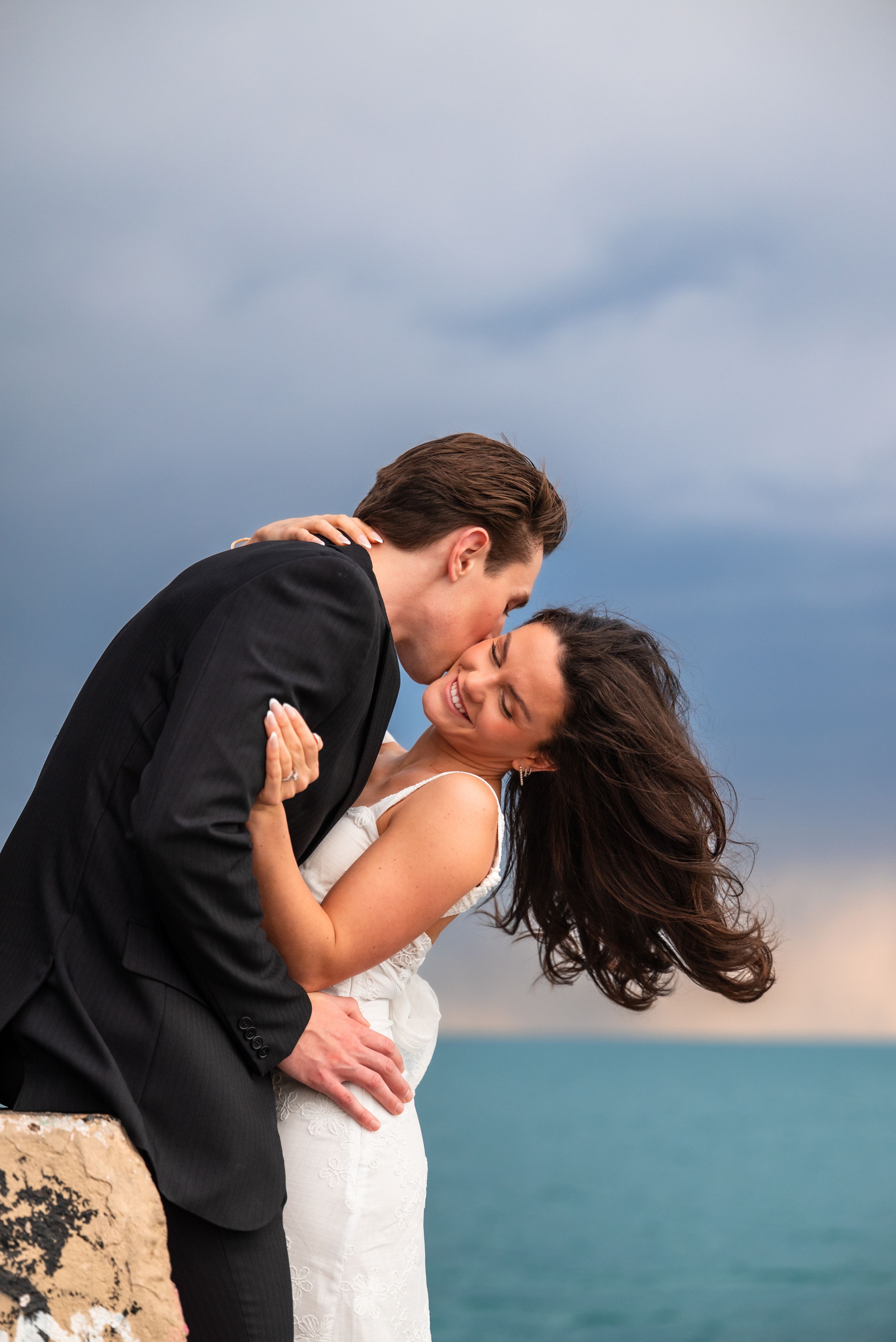 Newlywed couple embracing by the ocean, groom in black suit kissing bride in white dress with stormy sky backdrop.