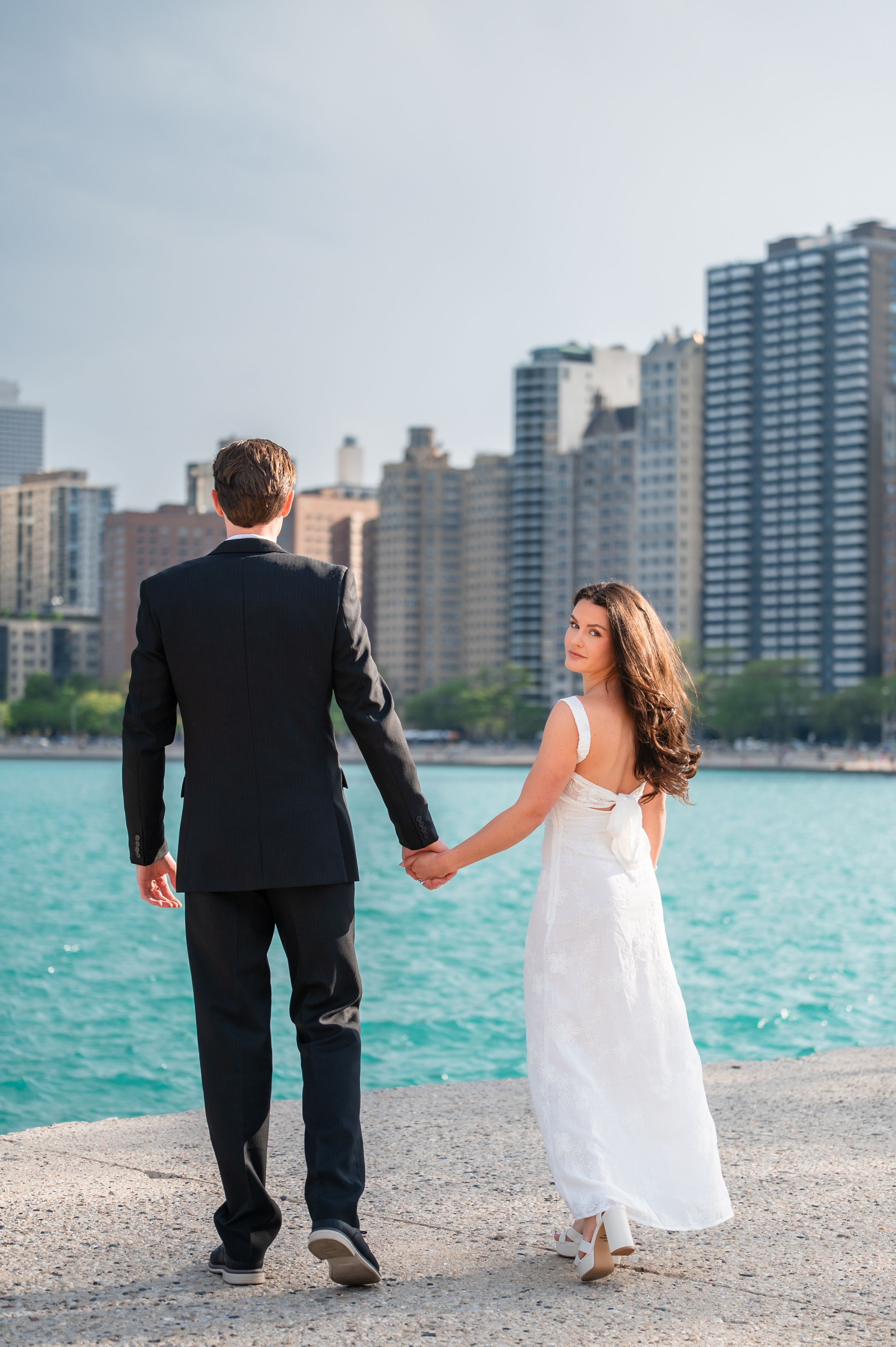 Couple in wedding attire holding hands by turquoise waterfront with Chicago skyline in background.