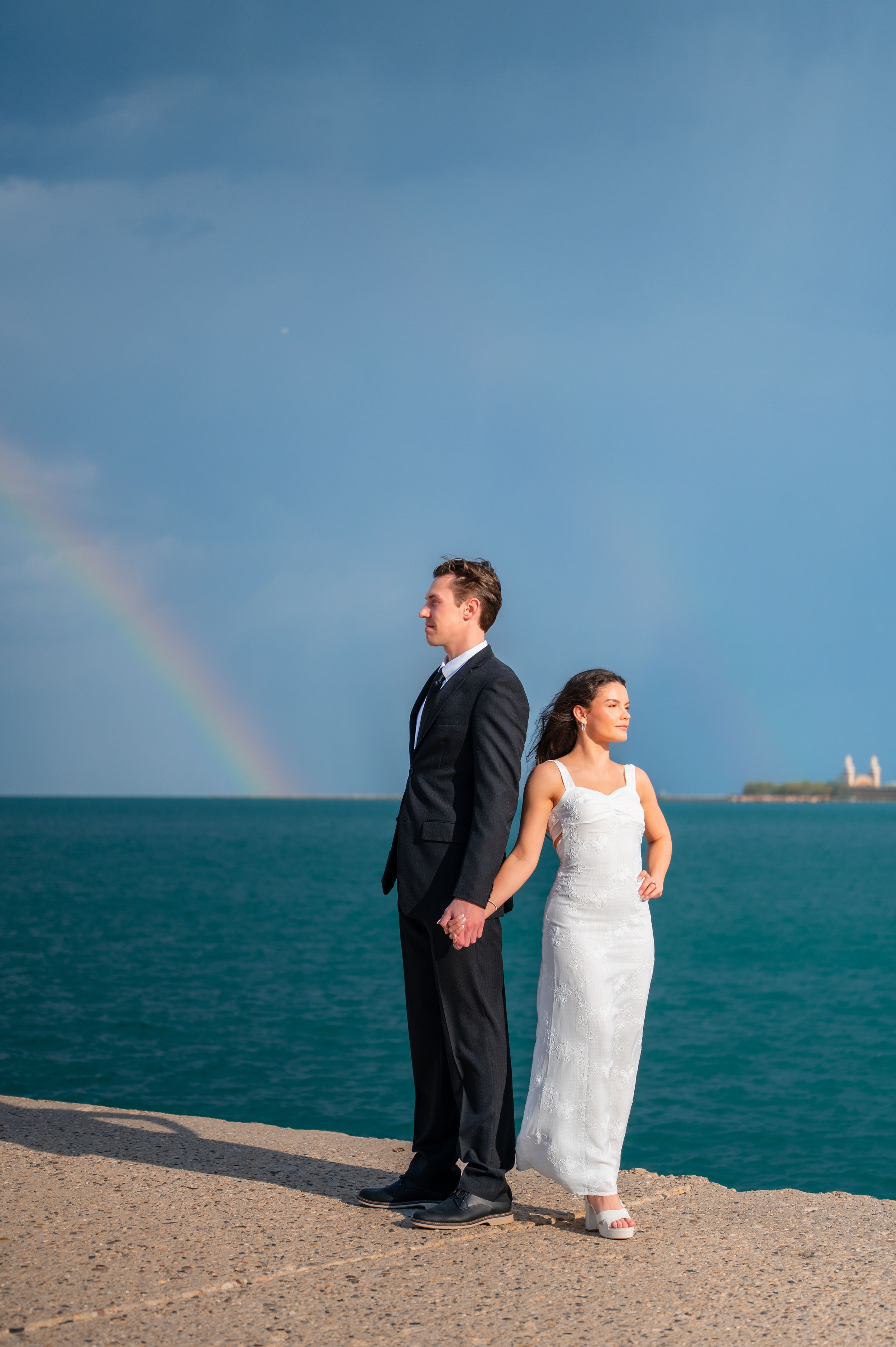 Couple in wedding attire holding hands on a waterfront pier with a rainbow over turquoise ocean waters.