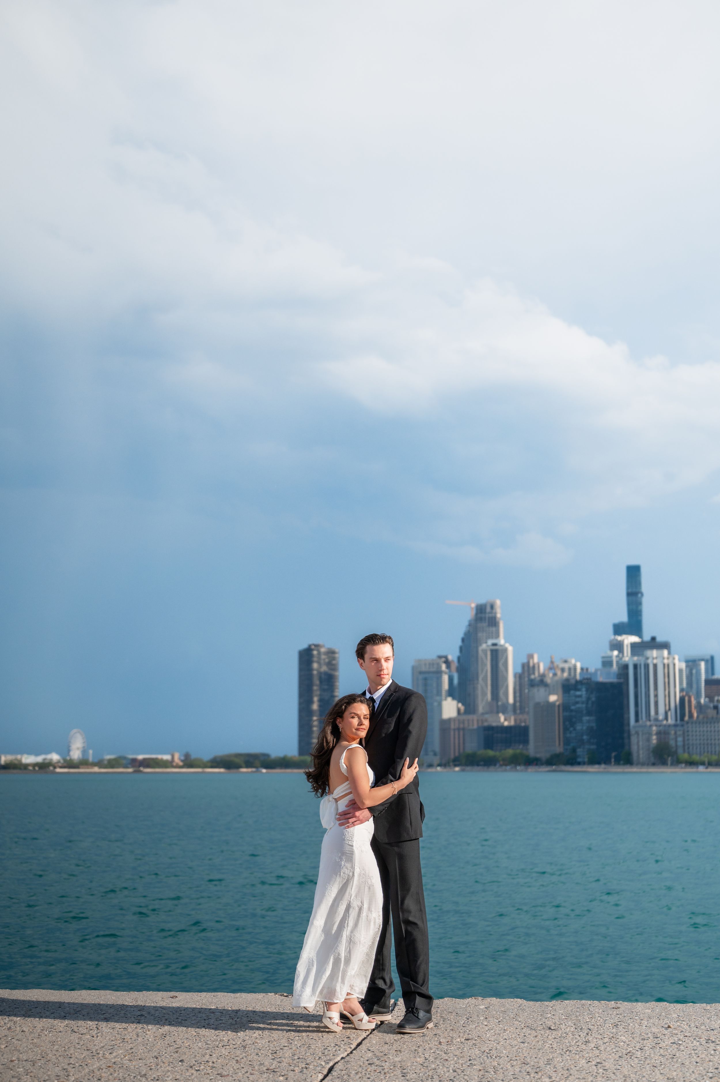 Couple embracing on Chicago lakefront with city skyline and dramatic cloudy sky in background.
