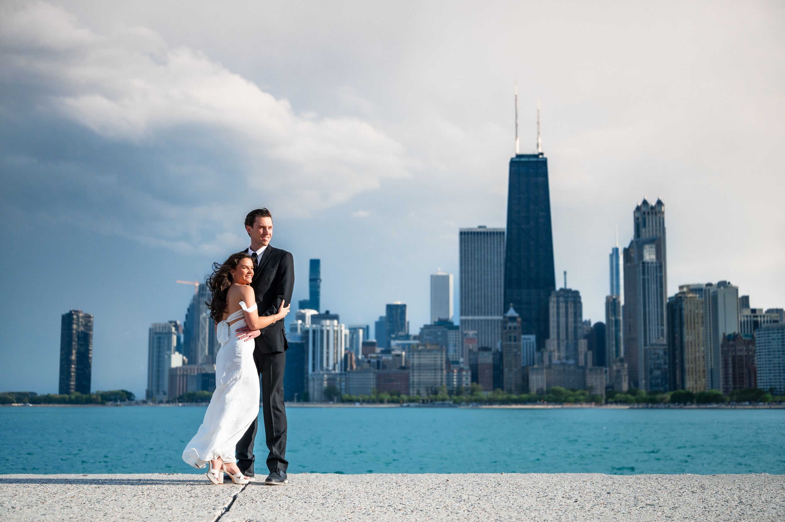 Bride and groom pose on Chicago lakefront with iconic city skyline and John Hancock Center in background.