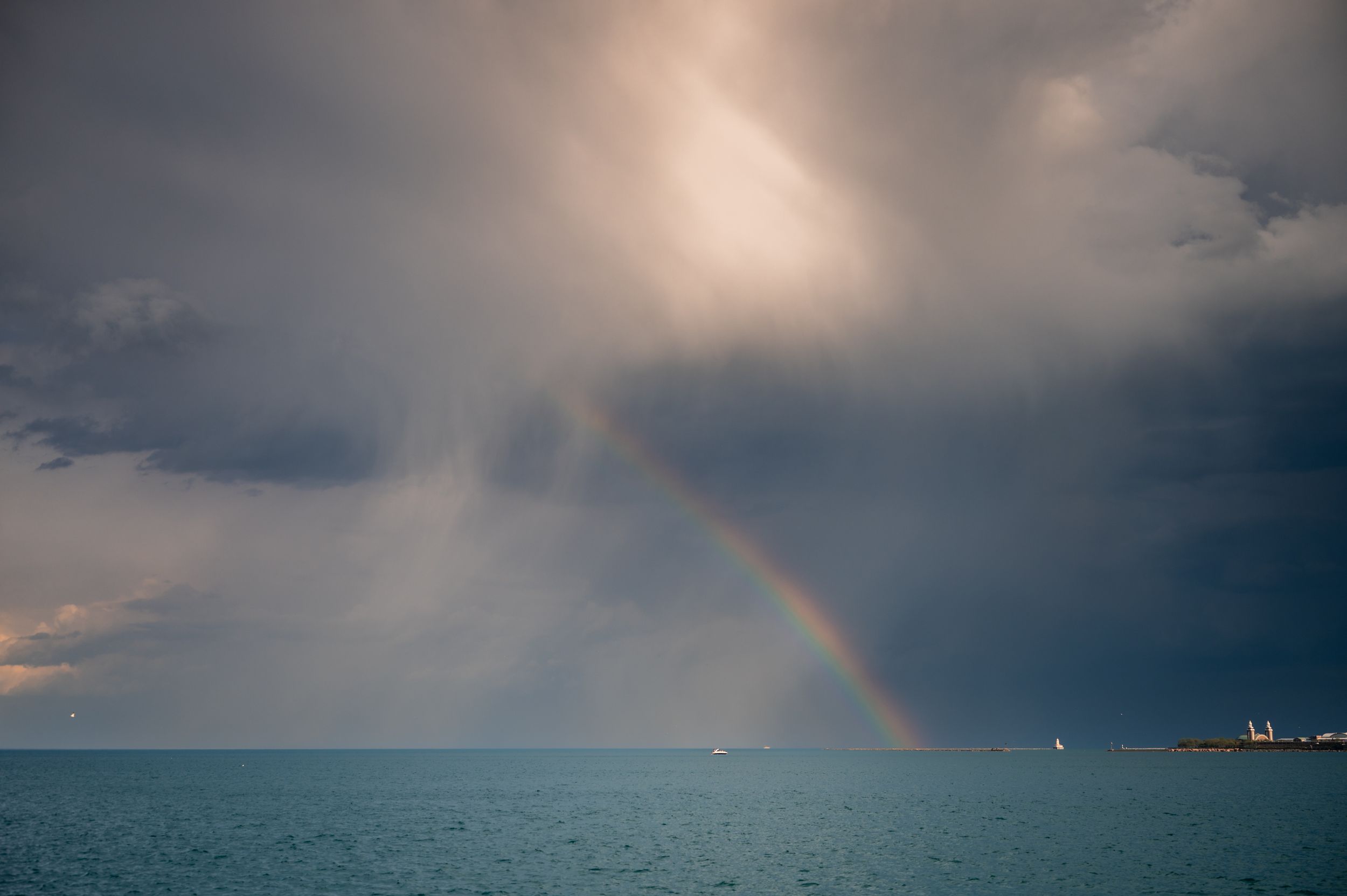 A vibrant rainbow arches over a calm turquoise sea beneath dramatic stormy grey clouds.