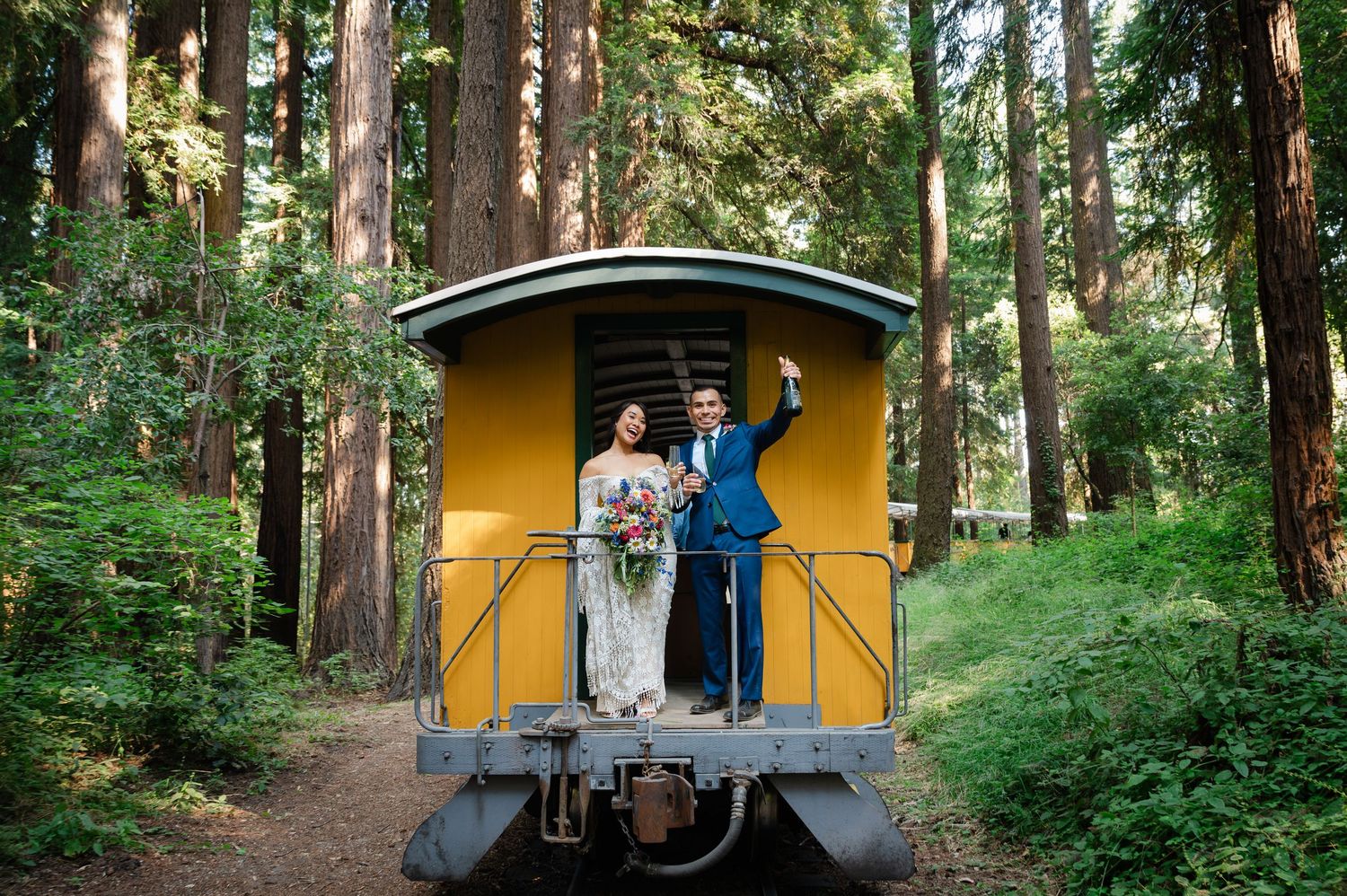 Bride and groom celebrating on a yellow vintage train car surrounded by tall redwood trees in a lush forest setting.