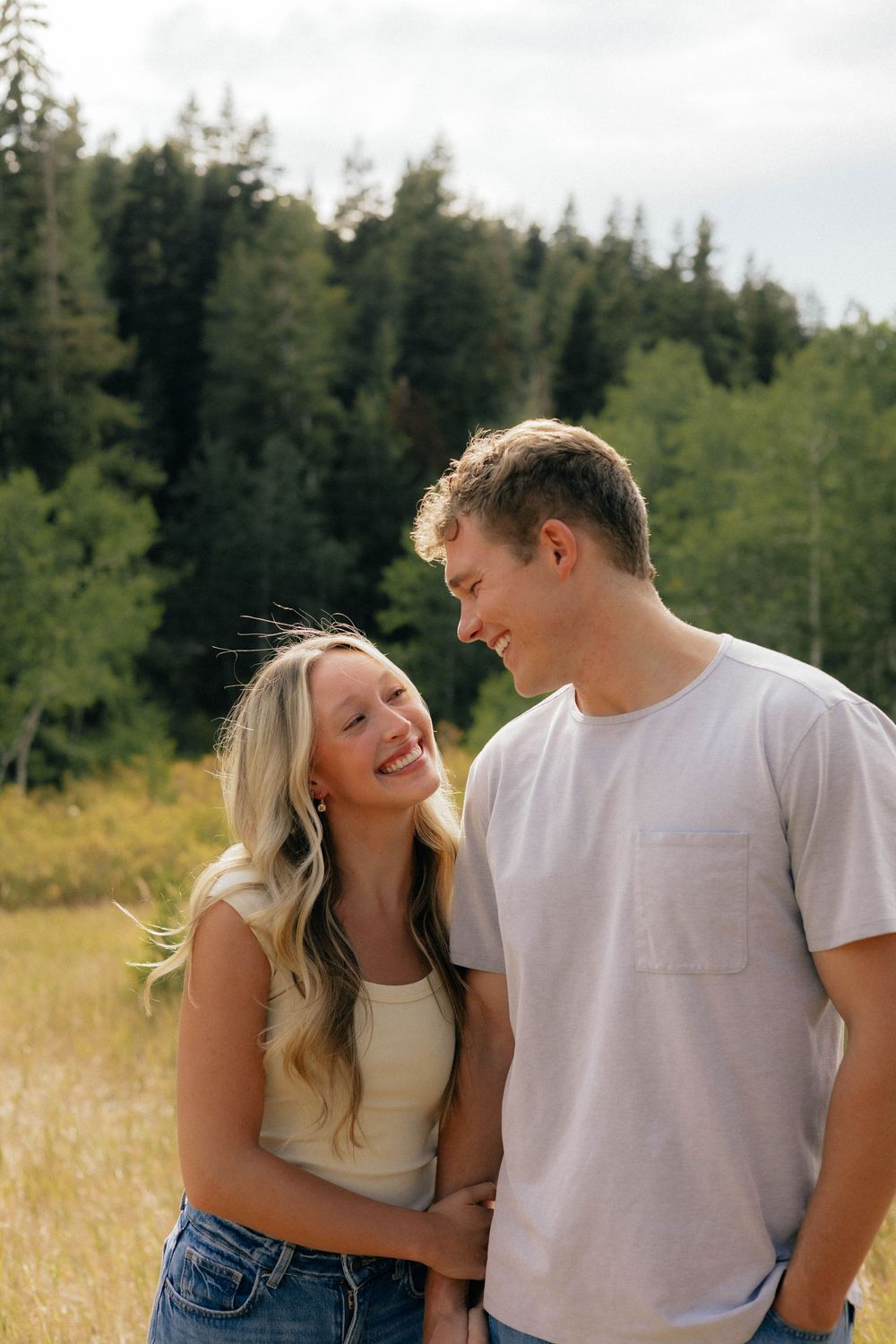 A smiling couple embraces in a sunny meadow with lush green pine trees in the background.