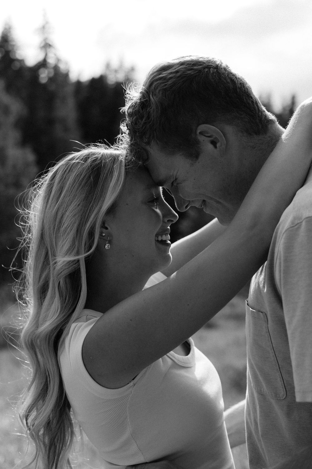 Black and white photo of a romantic couple leaning in for a kiss outdoors with trees in the background.
