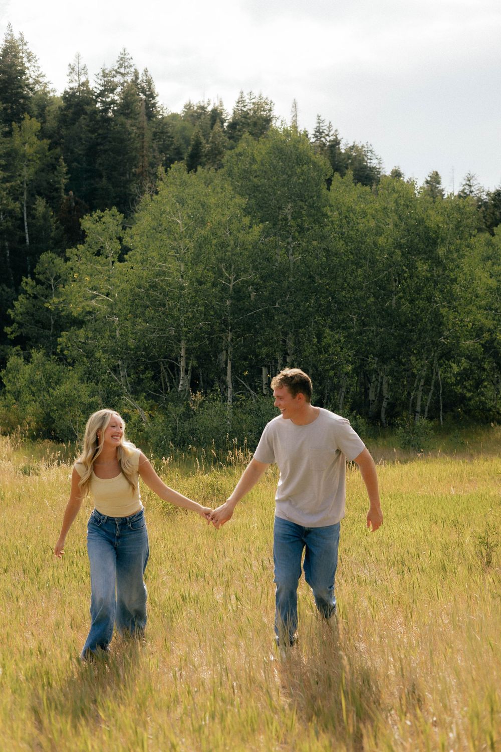 A couple holds hands while walking through a sunny meadow with lush green trees in the background.